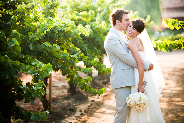 The happy couple standing in a vineyard - Bride is wearing white dress with full length veil and groom is wearing light blue suit - wedding portrait photo by Michael Norwood Photography